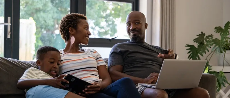 A smiling family of three sits on a dark sofa in a bright living room, with the father using a laptop, the mother looking toward him, and their young son playing on a tablet.